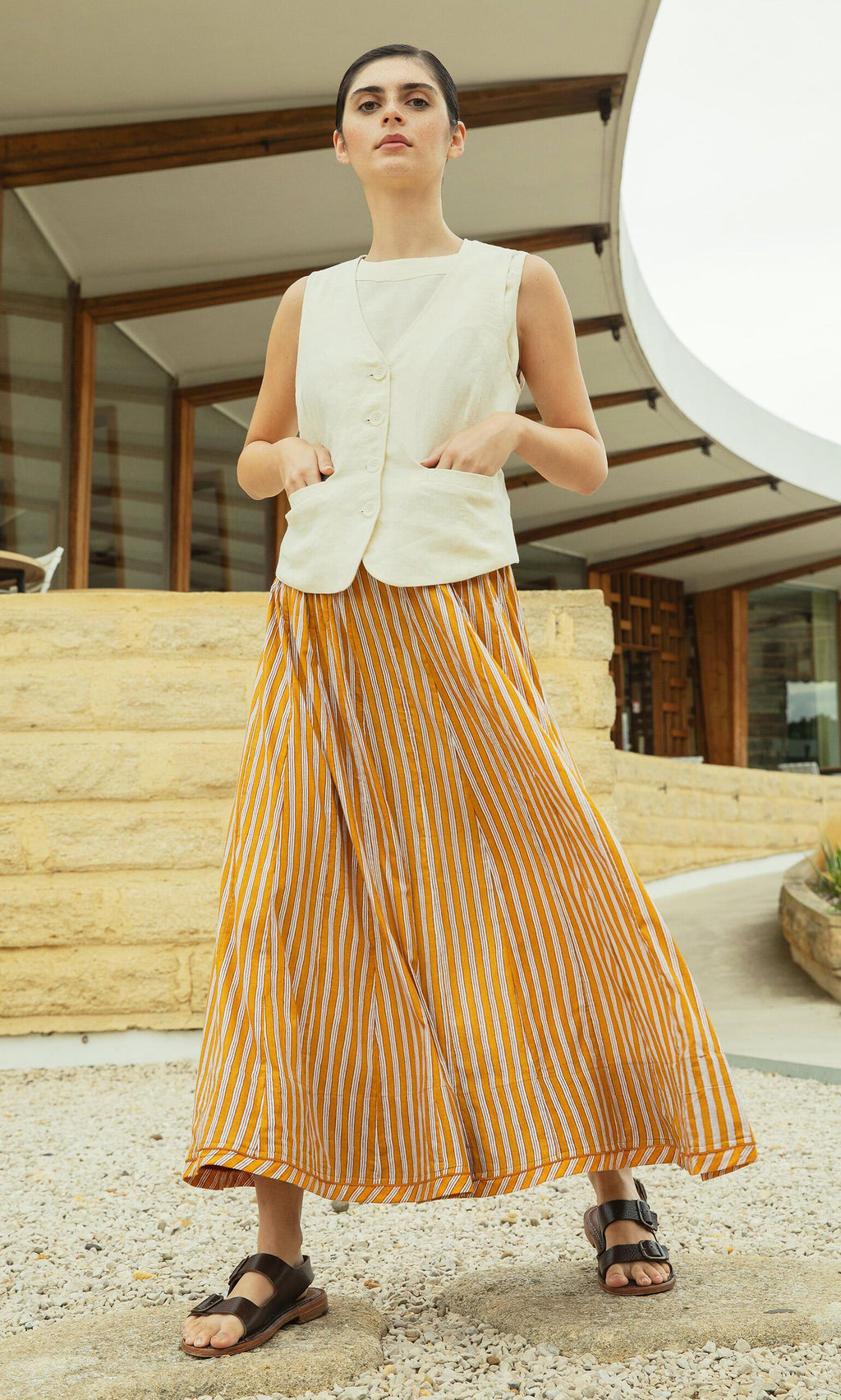 Plümo- Woman in a white vest and mustard skirt stands outside a modern building on a gravel path.