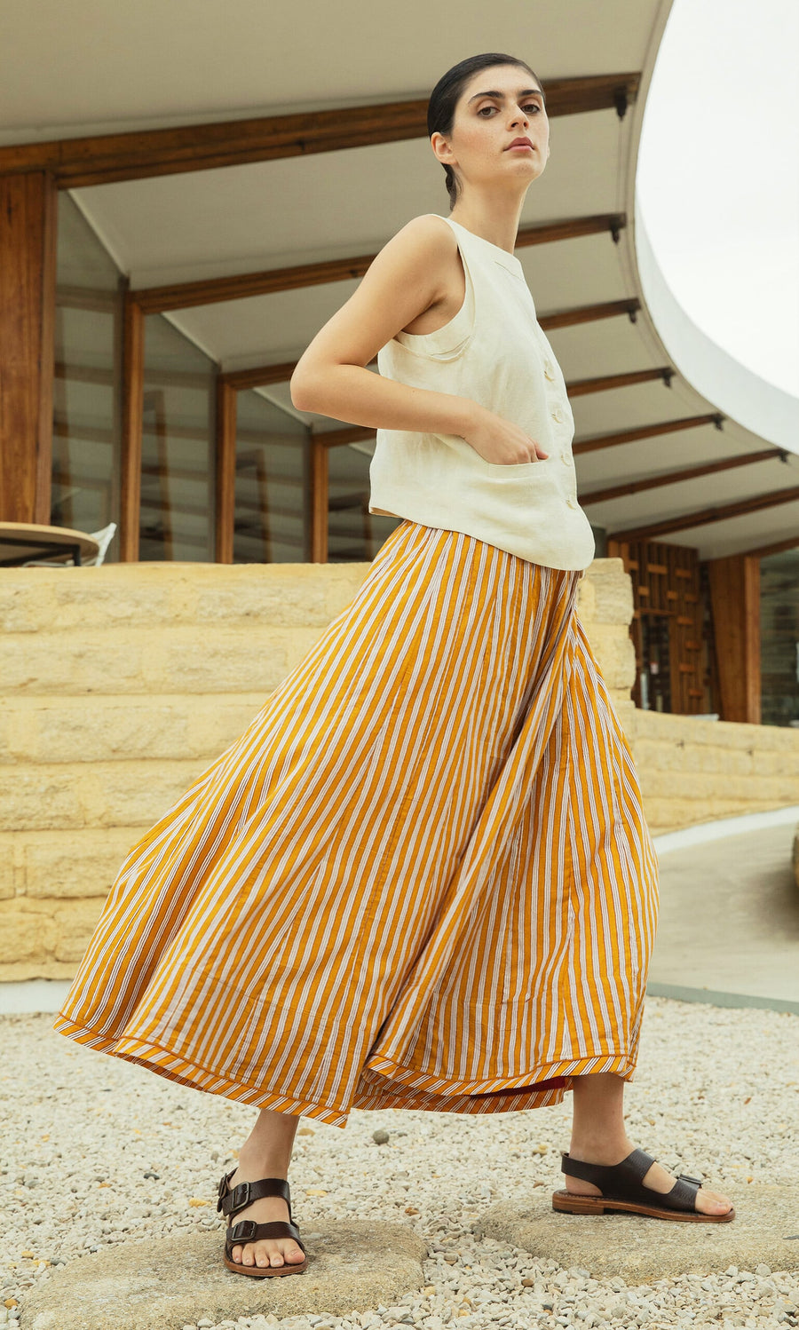 Plümo- Woman in a sleeveless white top and Mustard Skirt walking outdoors near a modern building.