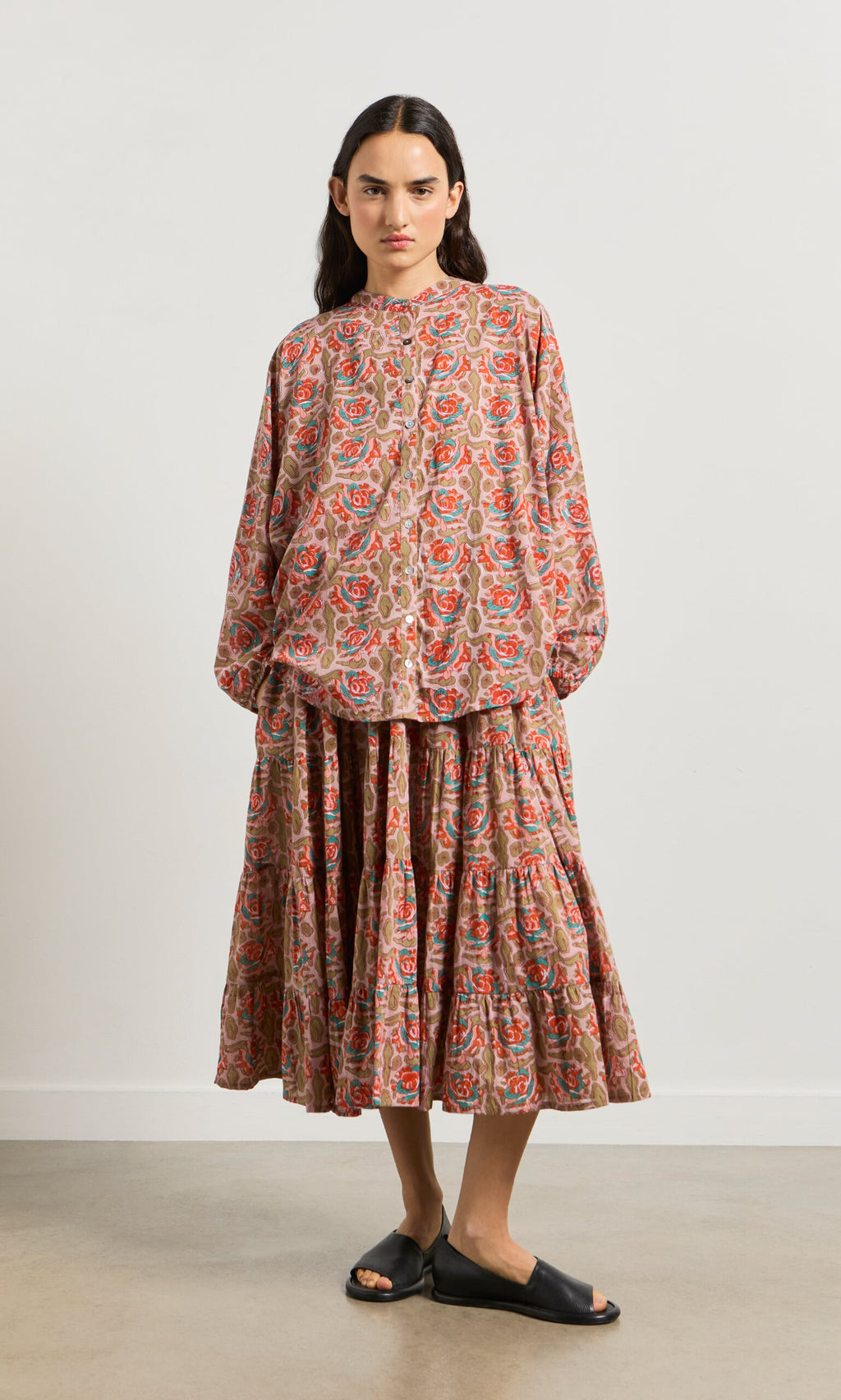 Plümo- Woman wears the Nila Flower Dress, a loose block-printed floral style, and black slide sandals against a plain wall.