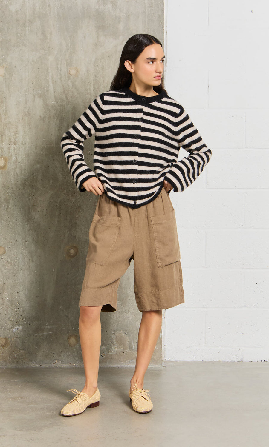 Plümo- Person in a black and white striped sweater and brown linen shorts stands against a concrete wall.