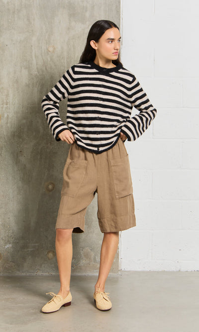 Plümo- Wearing the Nola Stripe Cardigan, tan knee-length shorts, and beige shoes, a person stands by a concrete wall.