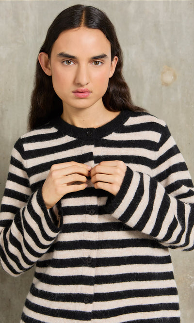 Plümo- Woman in a Nola Stripe Cardigan with long dark hair stands against a concrete wall.