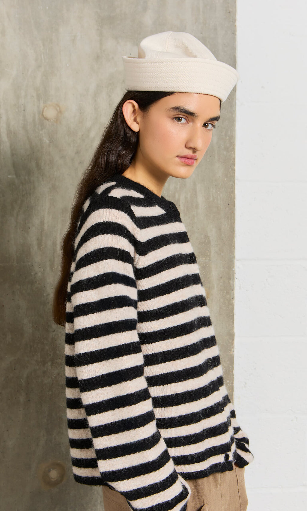 Plümo- Young woman in a Nola Stripe Cardigan and white hat stands by a concrete wall, looking at the camera.