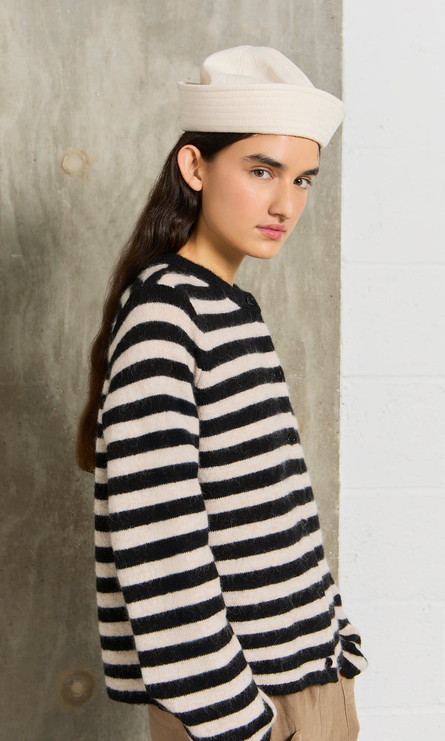 Plümo- Young woman in a Nola Stripe Cardigan and white hat stands by a concrete wall, looking at the camera.