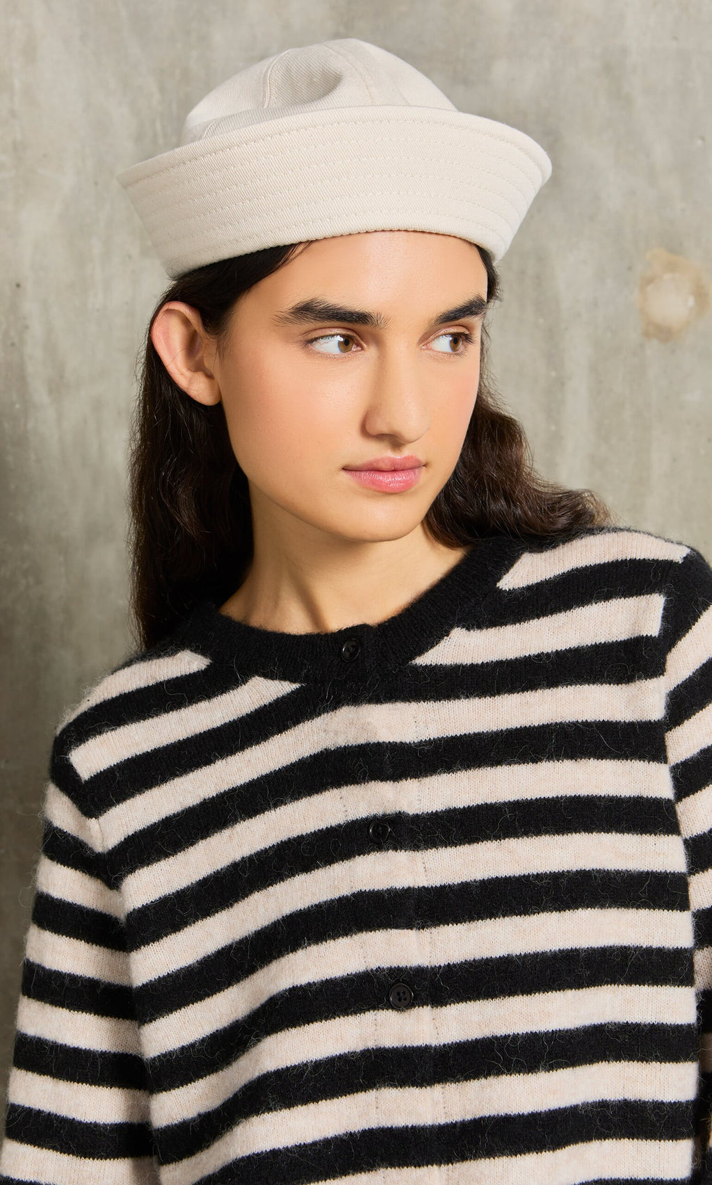 Plümo- Woman in the Nola Stripe Cardigan and white hat looks to the side, standing against a gray concrete wall.