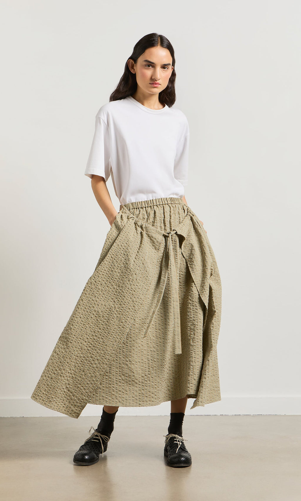 Plümo- Woman in a white tee and Nori Wrap Skirt, hands in pockets, standing on a light-colored floor.