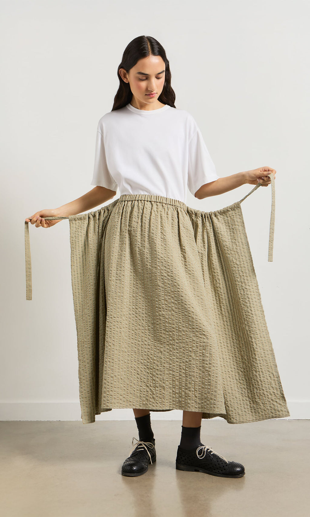 Plümo- Woman in white shirt and Nori Wrap Skirt holding two ties, standing on a gray floor.