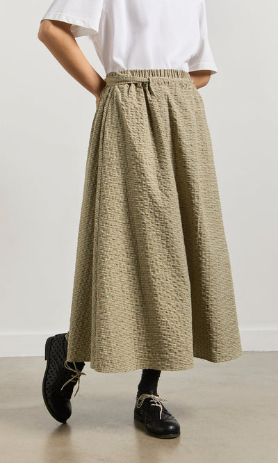 Plümo- Person wears a white shirt, Nori Wrap Skirt in tan gingham, black socks, and black shoes on a gray floor indoors.