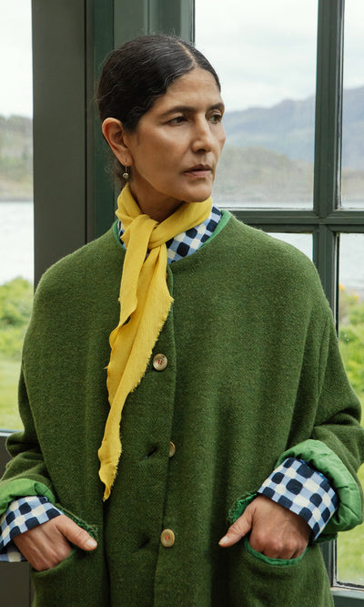 Plümo  Woman in green coat and checkered shirt wears the Norma Necktie, standing by a window, looking to the side.