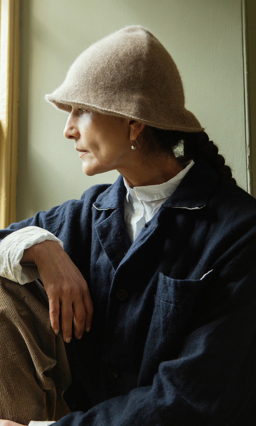 Plümo  Woman in a Felted Hat and dark coat sits by a window, gazing outside with a thoughtful expression.