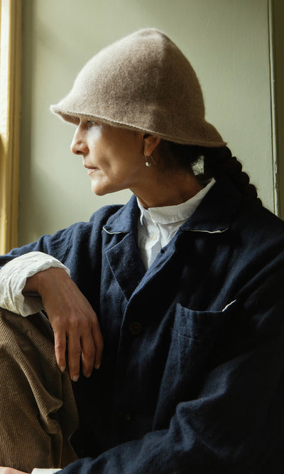 Plümo  Woman in a felt hat and Dunbar Tweed Jacket sits by a window, gazing outside with a thoughtful look.