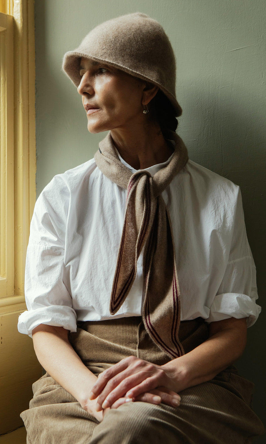 Plümo  Woman in a beige Felted Hat, scarf, white blouse, and brown skirt sits by a window, looking thoughtfully aside.