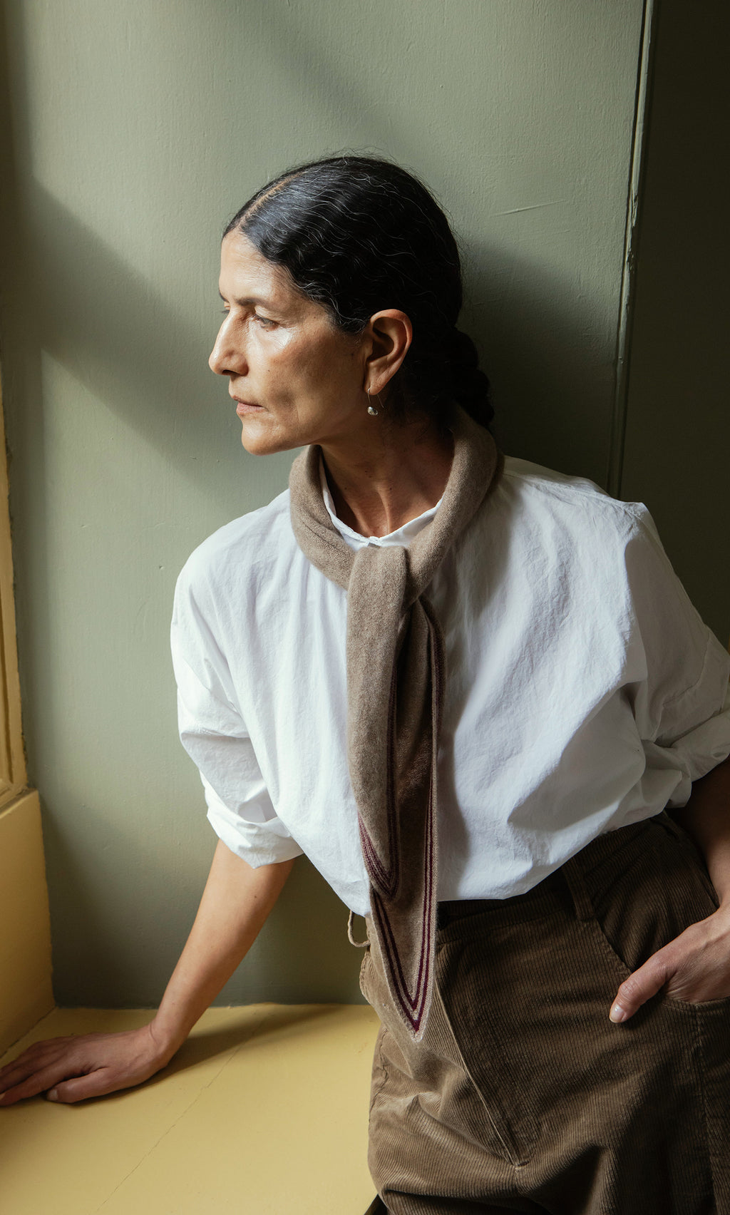 Plümo  Woman in a white shirt and Oatmeal Necktie sits by a window, gazing outside with a thoughtful expression.