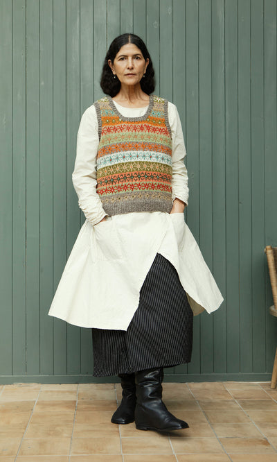 Plümo  Woman in Oban Fairisle Top, white dress, striped skirt, and black boots stands by a green wall.