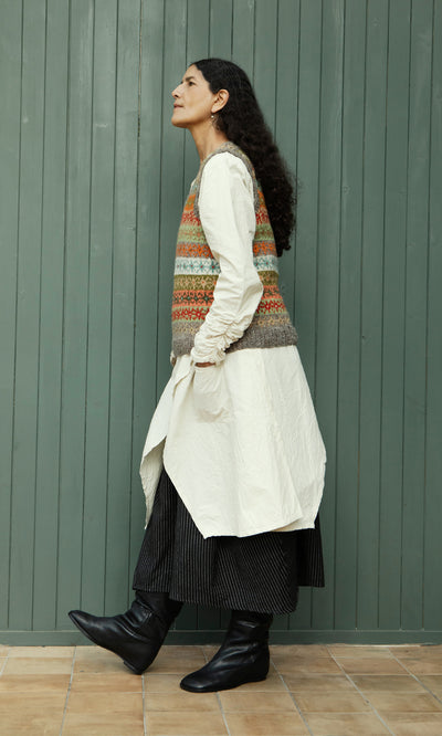 Plümo  Woman in profile wears a long white dress and the Oban Fairisle Top, long black skirt, and black boots indoors.