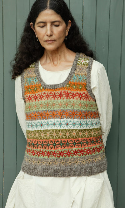 Plümo  Woman wearing the Oban Fairisle Top and white dress stands, eyes closed, in peaceful reflection by green doors.