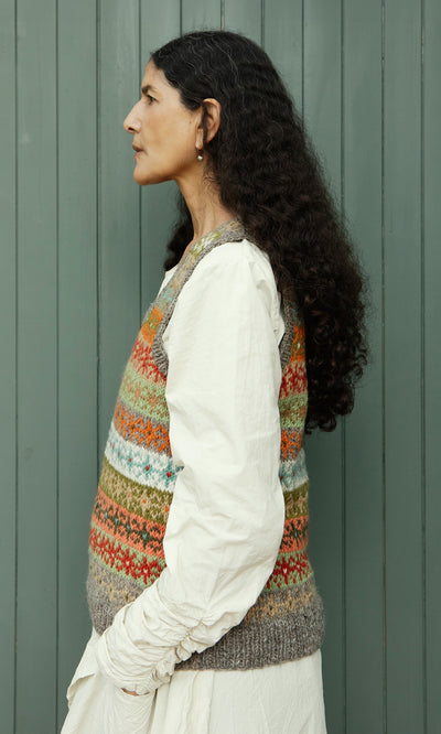 Plümo  Woman in profile wears the Oban Fairisle Top over a cream dress, standing by green wooden panels.