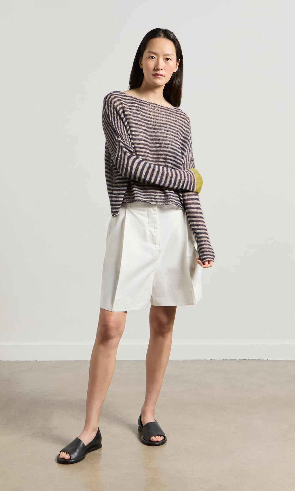 Plümo- Woman in the Paloma Stripe Sweater, white shorts, and black sandals stands on a concrete floor.