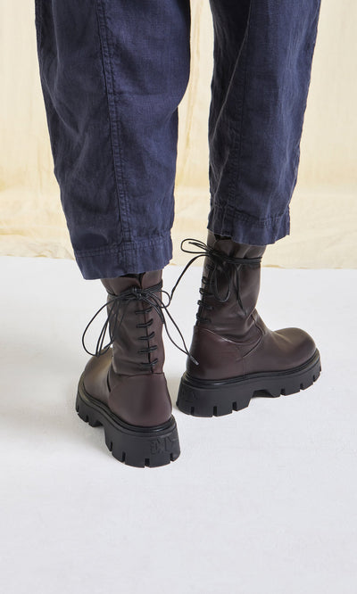 Plümo- Person in loose dark blue pants wearing Paola Boots—artisan-crafted brown lace-up boots with thick black soles.