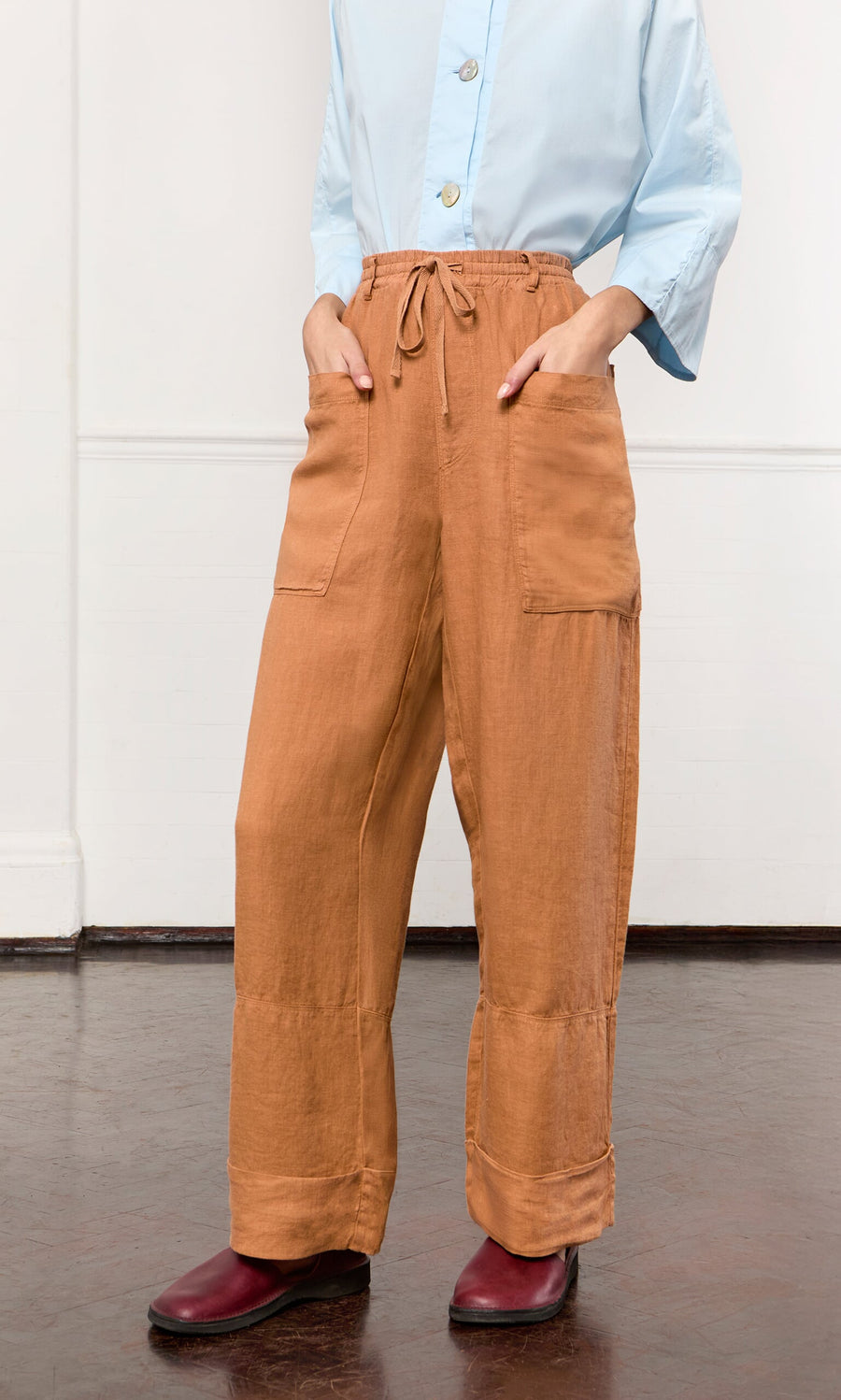 Plümo- Person in Pecan Pocket Pants, light blue shirt, and red shoes stands against a white wall.