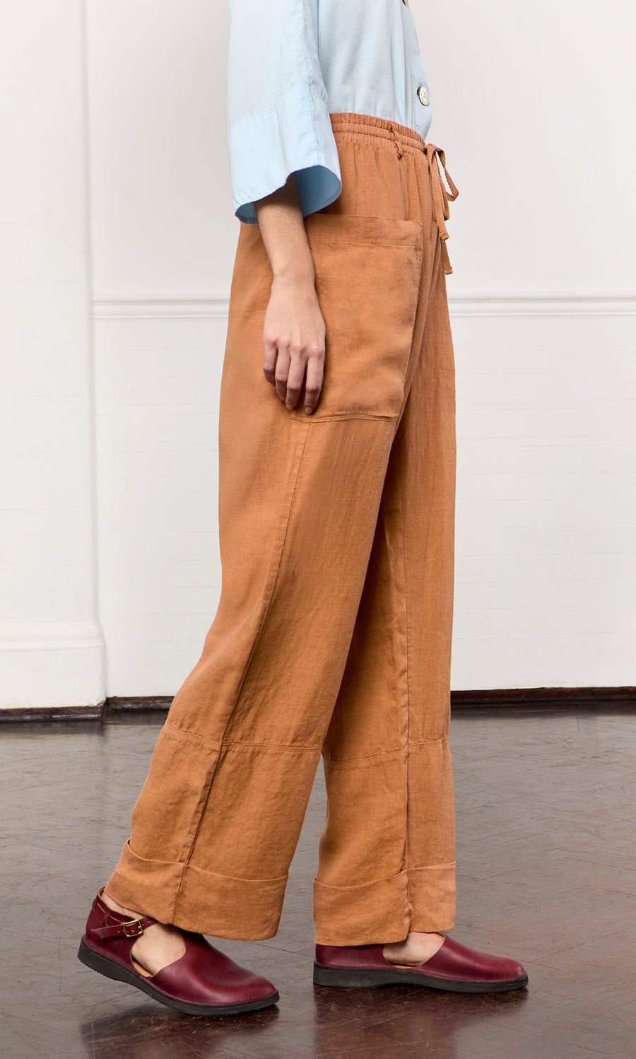 Plümo- Person indoors wearing Pecan Pocket Pants in tan with a drawstring waist, a light blue top, and dark red flats.