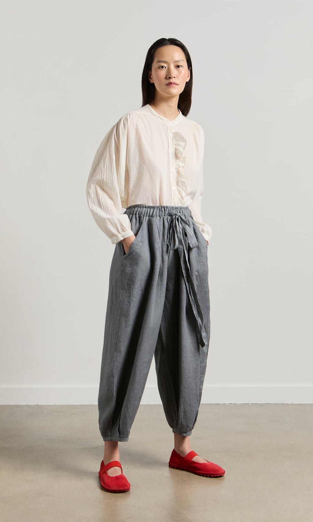 Plümo- Woman indoors wearing Percival Linen Pants in gray with balloon legs, elastic waist, and pockets, paired with a white blouse.