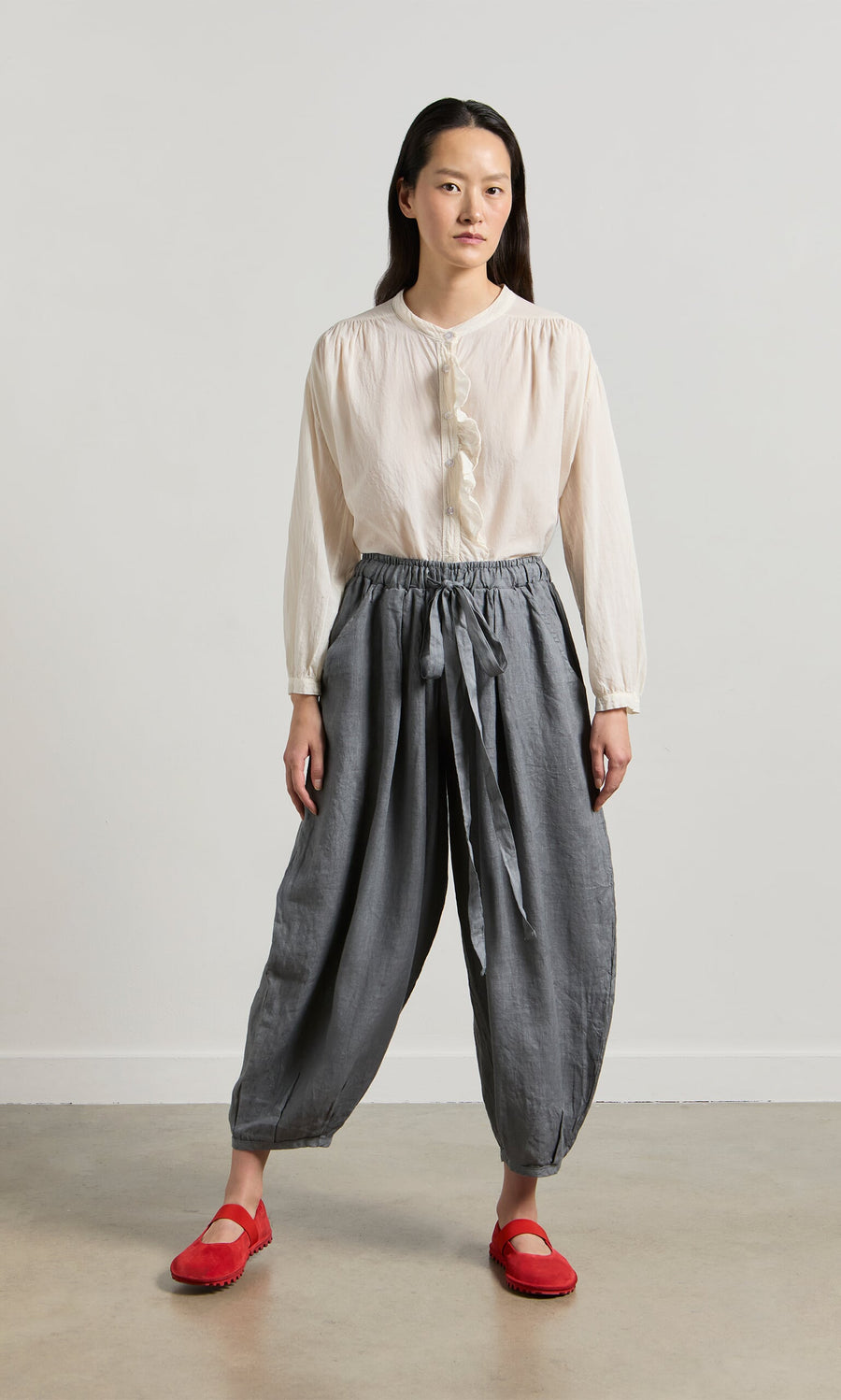 Plümo- A woman wears Percival Linen Pants with an elastic waist, a white blouse, and red shoes against a plain background.