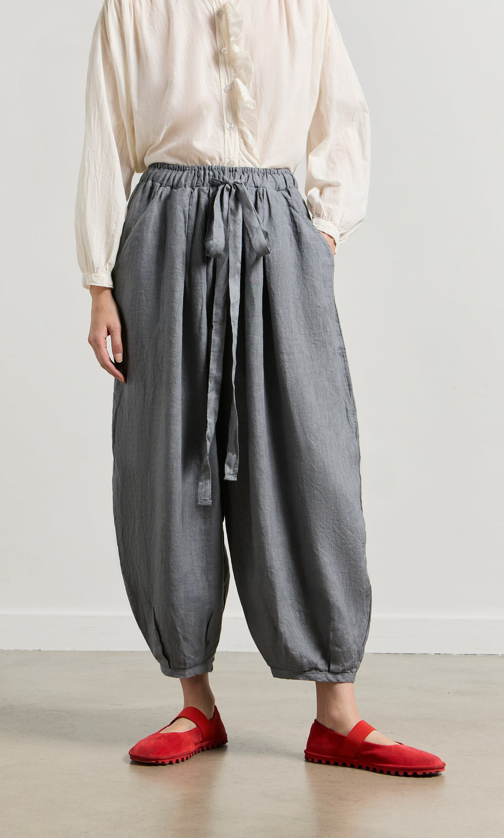 Plümo- Person wearing Percival Linen Pants, a tie-waist gray style, with an off-white blouse and red shoes, standing indoors.