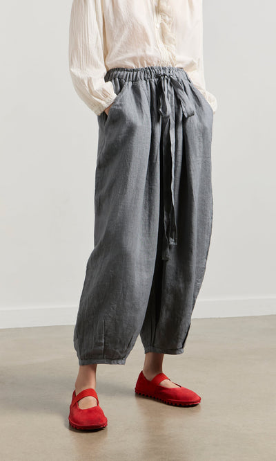 Plümo- Person wearing Percival Linen Pants with an elastic waist, a white blouse, and bright red shoes standing indoors.