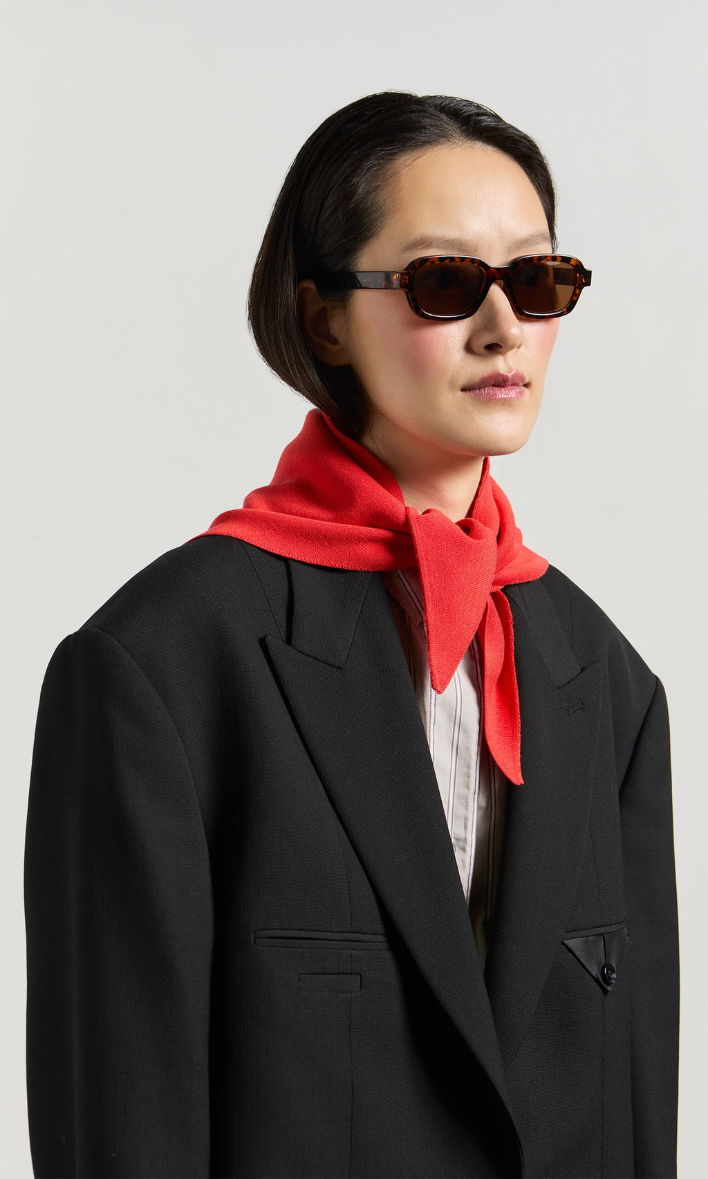 Plümo- Person in Portia Sunglasses and black blazer with a red sweater draped around neck, against a plain background.