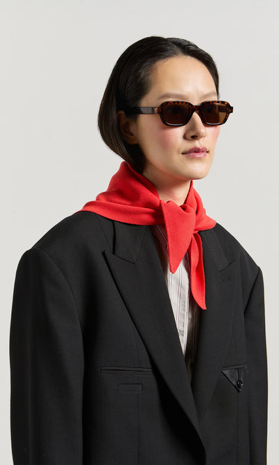 Plümo- Person in Portia Sunglasses and black blazer with a red sweater draped around neck, against a plain background.