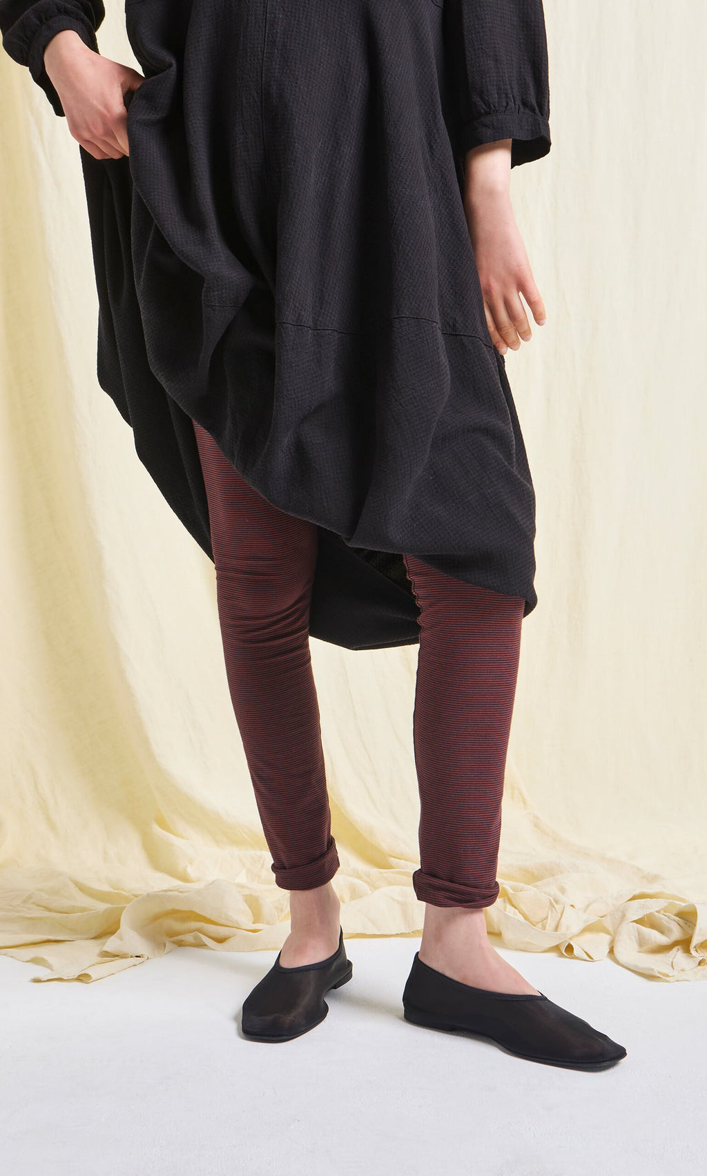Plümo- Person wearing Prue Leggings in maroon cotton with a black dress and flats, standing on a cream backdrop.