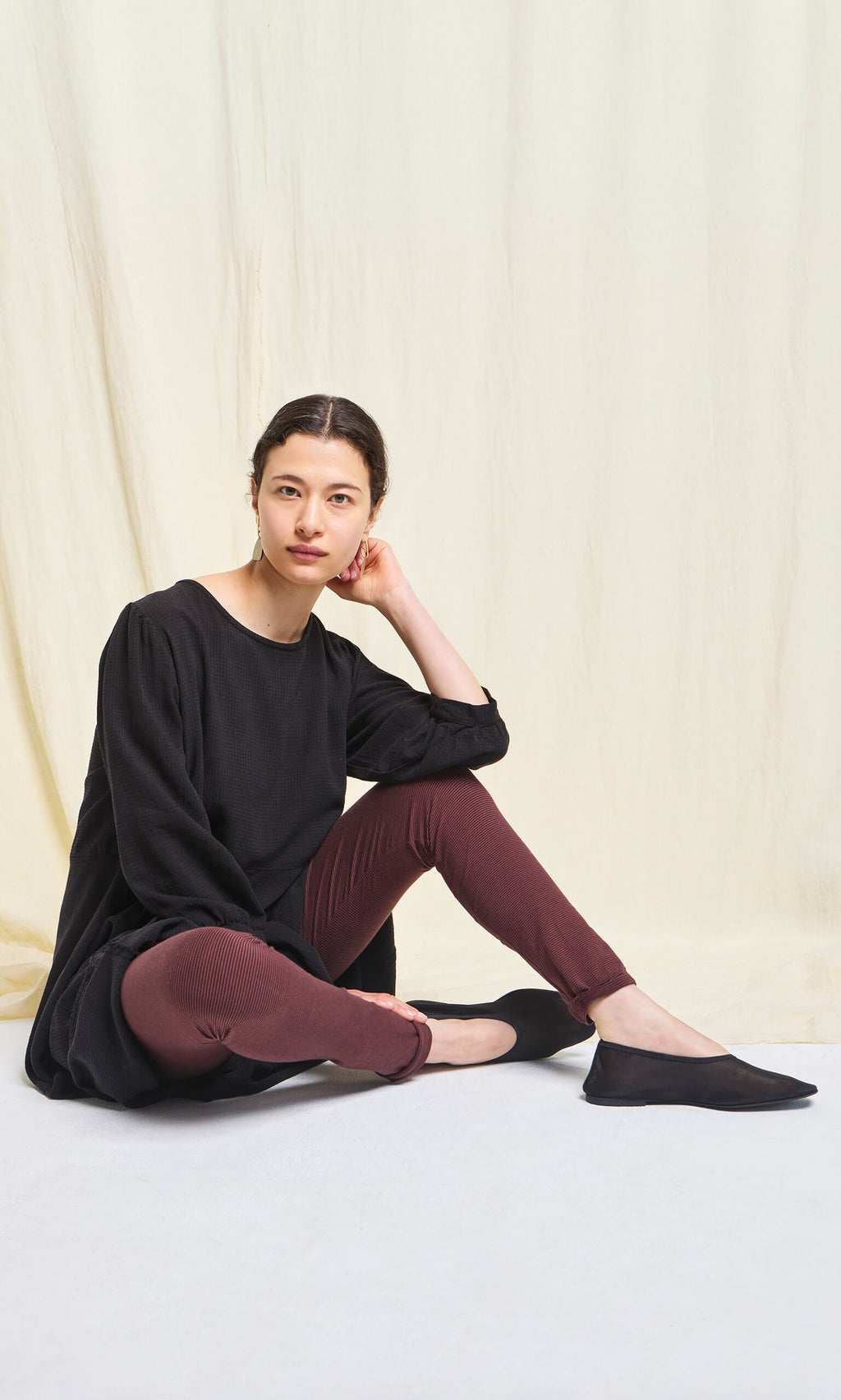 Plümo- Woman seated on the floor in Prue Leggings and a black top, wearing black flats against a neutral background.