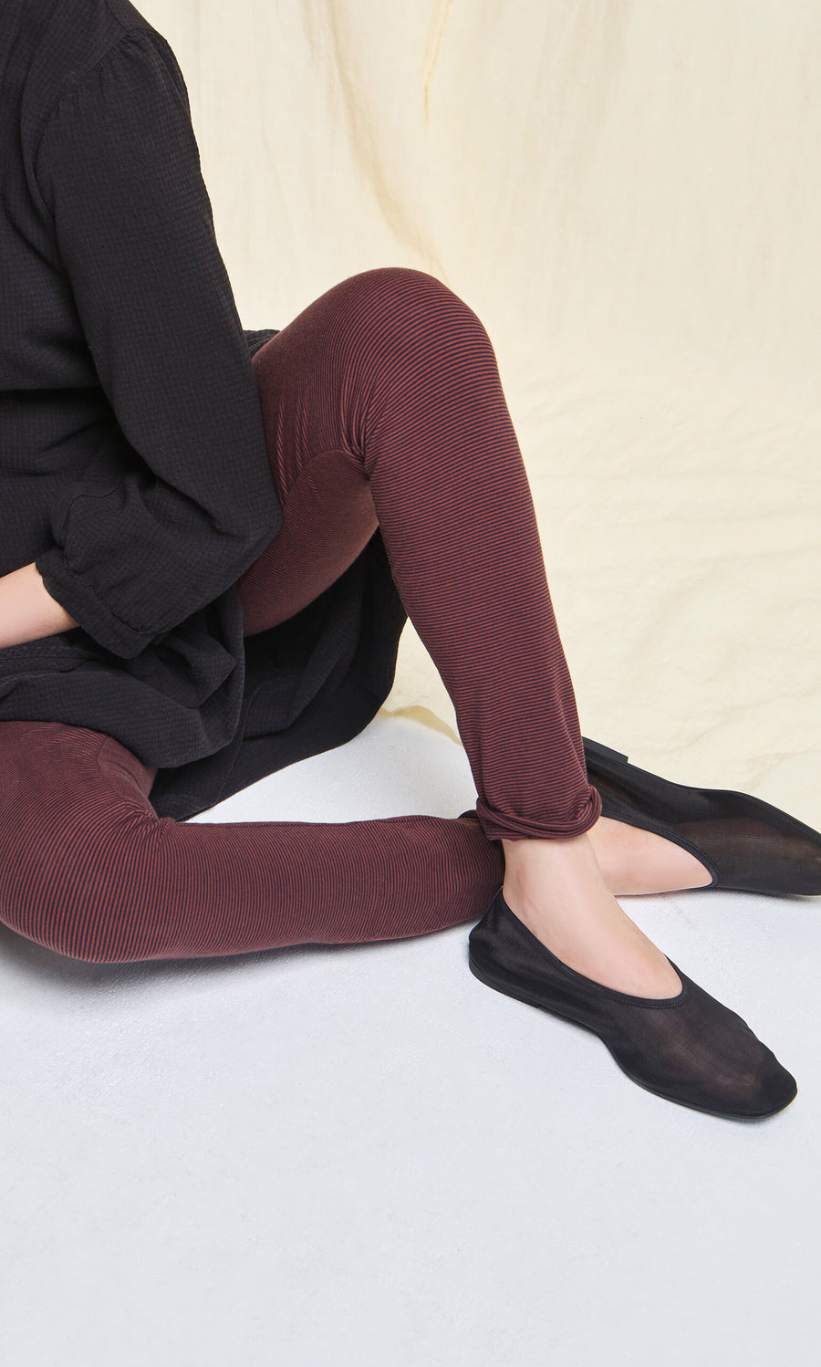 Plümo- Person seated on the floor wearing maroon Prue Leggings, a black top, and black flat shoes against a light background.