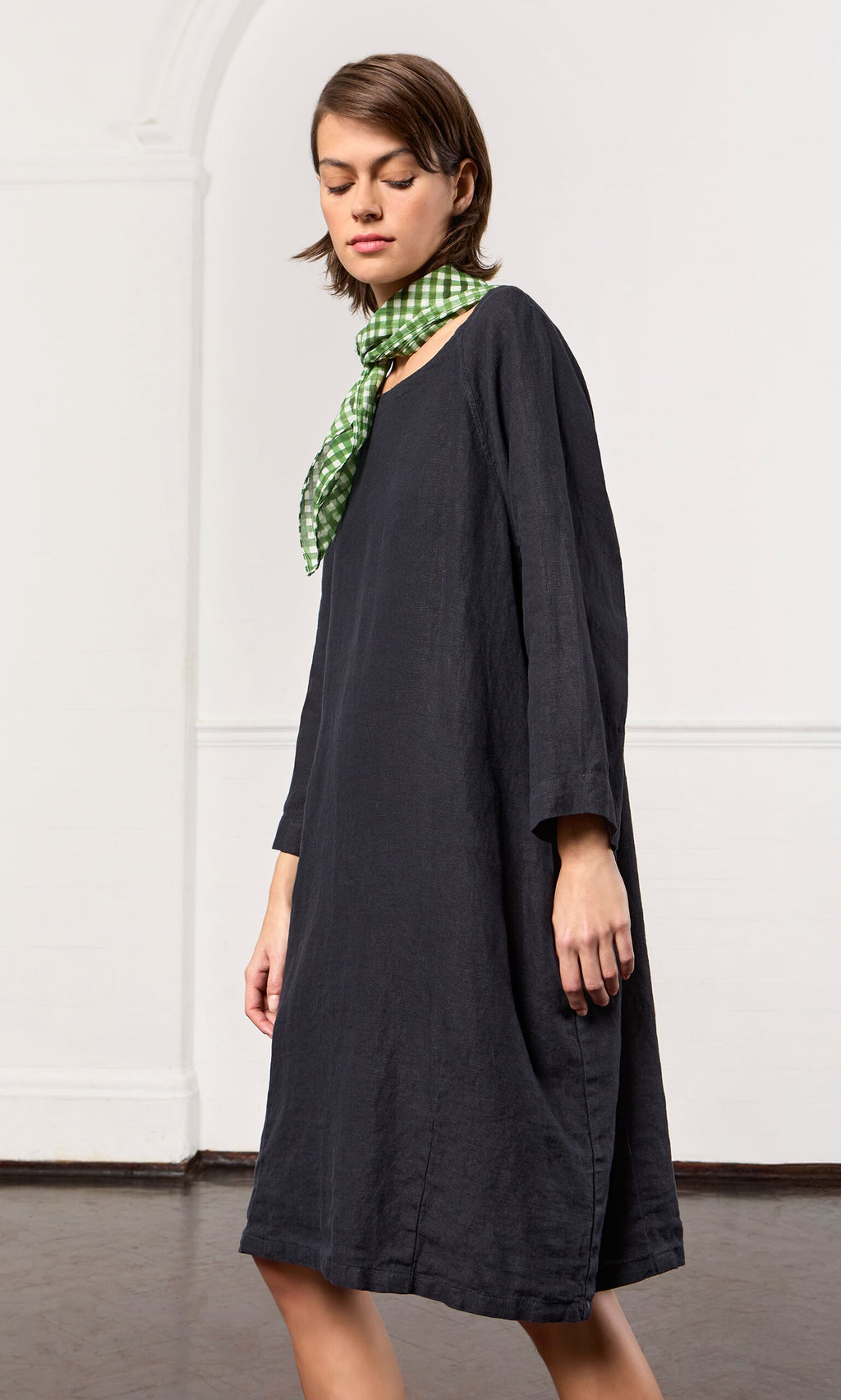 Plümo- Woman wearing a black Raglan Dress with bracelet sleeves and green-checkered scarf stands indoors by a white wall.