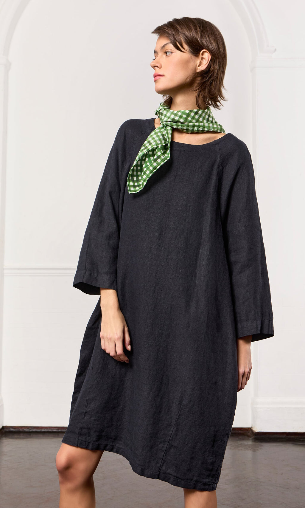 Plümo- Woman in a loose-fitting black Raglan Dress with bracelet-length sleeves and a green checkered scarf, indoors.