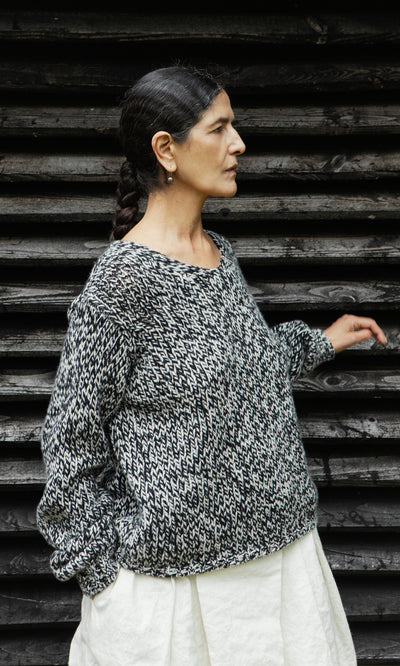 Plümo  Woman in a Rhian Wool Jumper and white skirt stands sideways against a dark wooden wall.