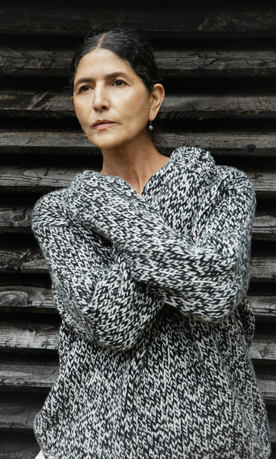 Plümo  Woman in a Rhian Wool Jumper stands with arms crossed by wooden slats, looking thoughtful and elegant.