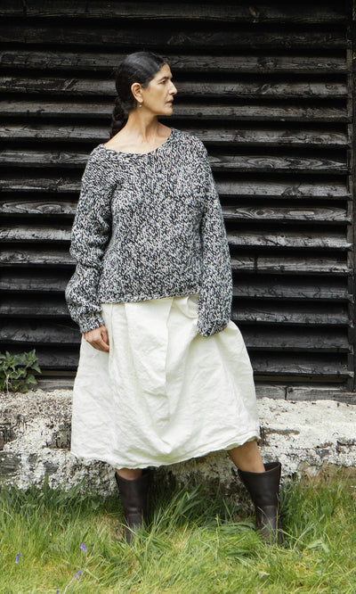 Plümo  Woman in Rhian Wool Jumper, white skirt, and brown boots stands by a wooden wall, looking to the side.