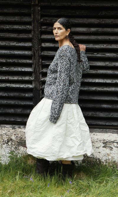 Plümo  Woman in a sustainable Rhian Wool Jumper stands on grass by a weathered, dark wooden wall.