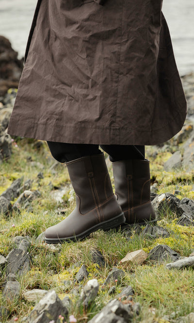 Plümo  Person in Rigger Boots and a long brown coat stands on grassy, rocky terrain beside the water.