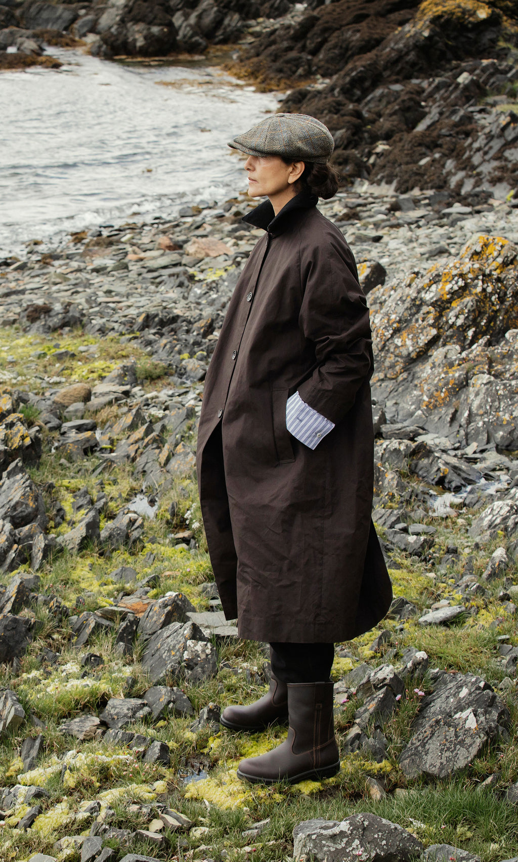 Plümo  Person in a brown coat, cap, and Rigger Boots stands on rocky grass by water, gazing into the distance.