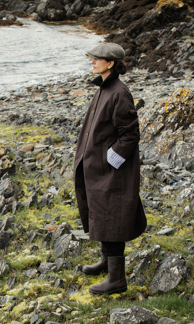 Plümo  Person in a brown coat, cap, and Rigger Boots stands on rocky grass by water, gazing into the distance.