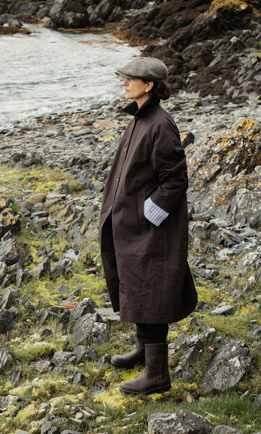 Plümo  Person in a brown coat, cap, and Rigger Boots stands on rocky grass by water, gazing into the distance.