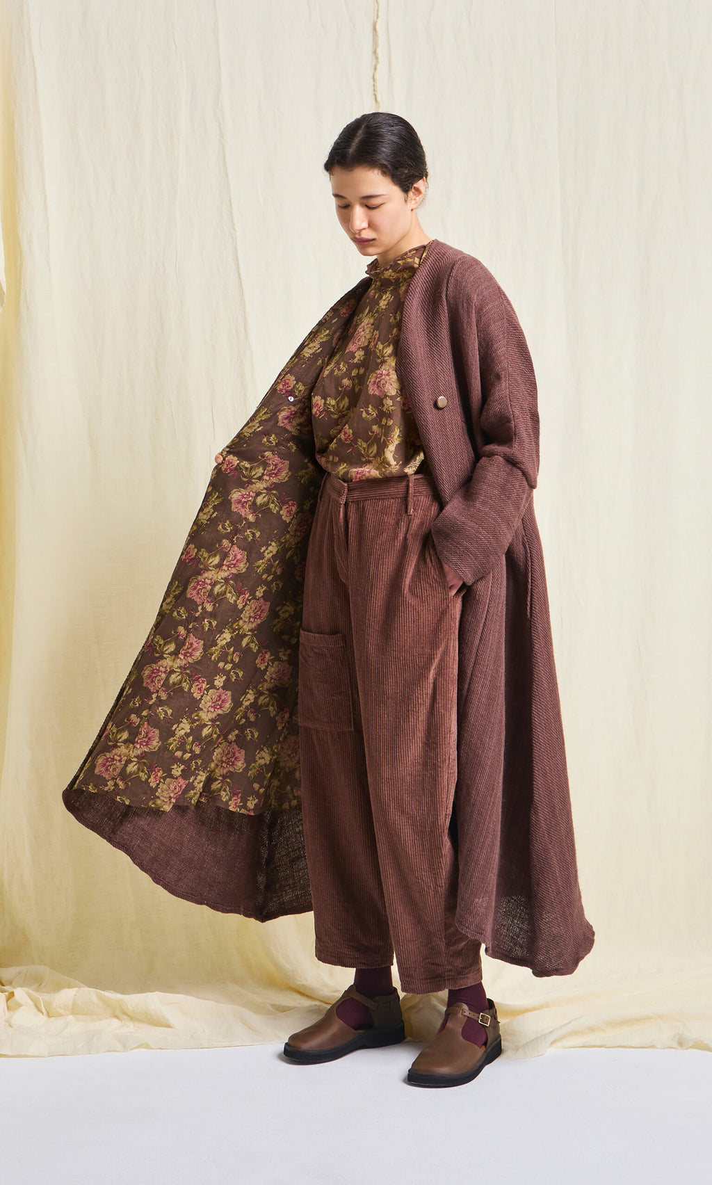 Plümo- A person in Lumber Pants, a brown floral top, and a long brown coat stands against a light fabric backdrop.