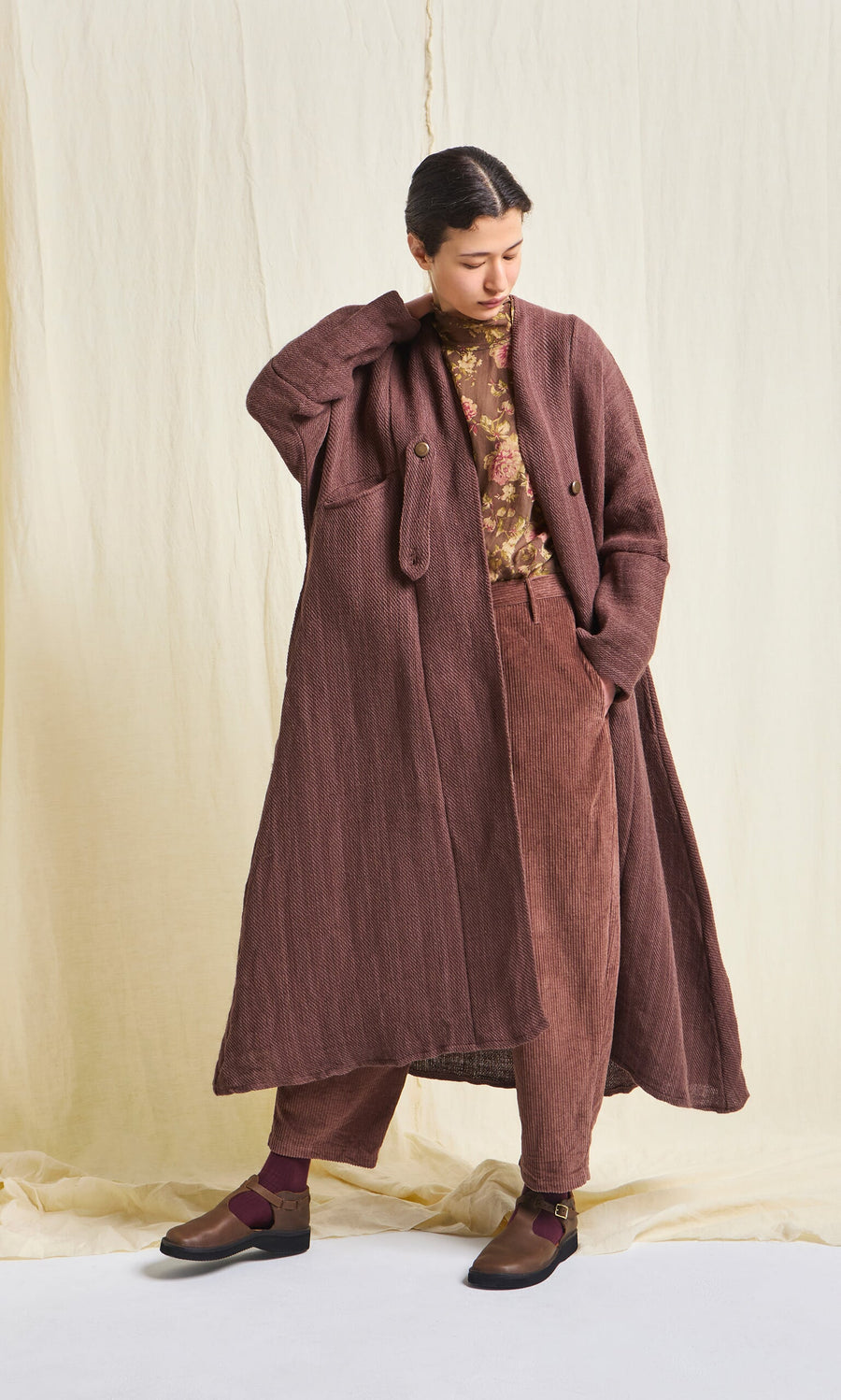 Plümo- Person wearing a Rosewood Coat, cotton floral print top, brown pants, and dark shoes stands by a light fabric background.