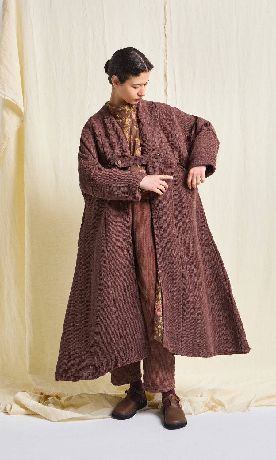 Plümo- A person in a Rosewood Coat with an A-line silhouette and brown pants stands against a cream fabric backdrop.