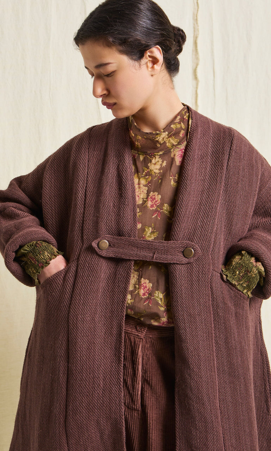Plümo- Woman wearing the Rosewood Coat over a cotton floral blouse, hands in pockets, looking down to her left.