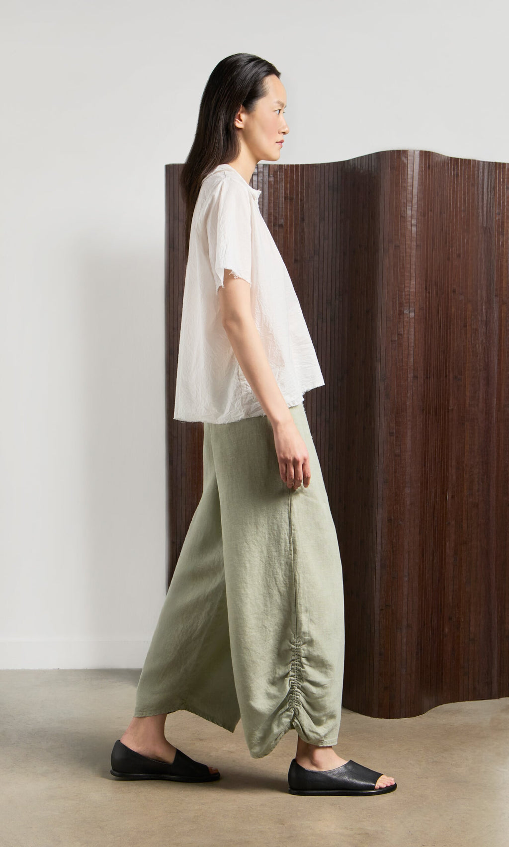 Plümo- A person in a white top, Royles Pants, and black sandals stands by a wooden room divider.