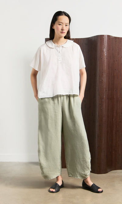 Plümo- Woman in a white blouse and Royles Pants with an elasticated waist stands before a brown folding screen.